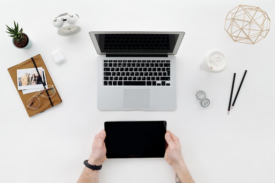 Bureau vu du dessus avec ordinateur portable, tablette tenue par deux mains et carnet, illustrant l'évaluation comparative d'outils numériques